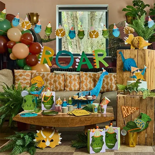 Dinosaur-themed party setup with ROAR banner, balloons, jungle decorations, and colorful tableware on a cosy indoor display